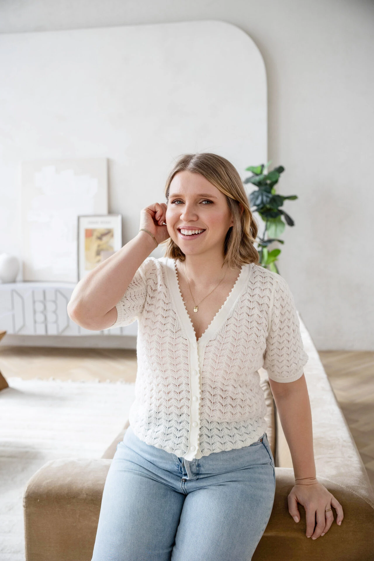 Smiling woman in a white knit cardigan sits on a beige couch in a bright living room with plants and frames in the background.