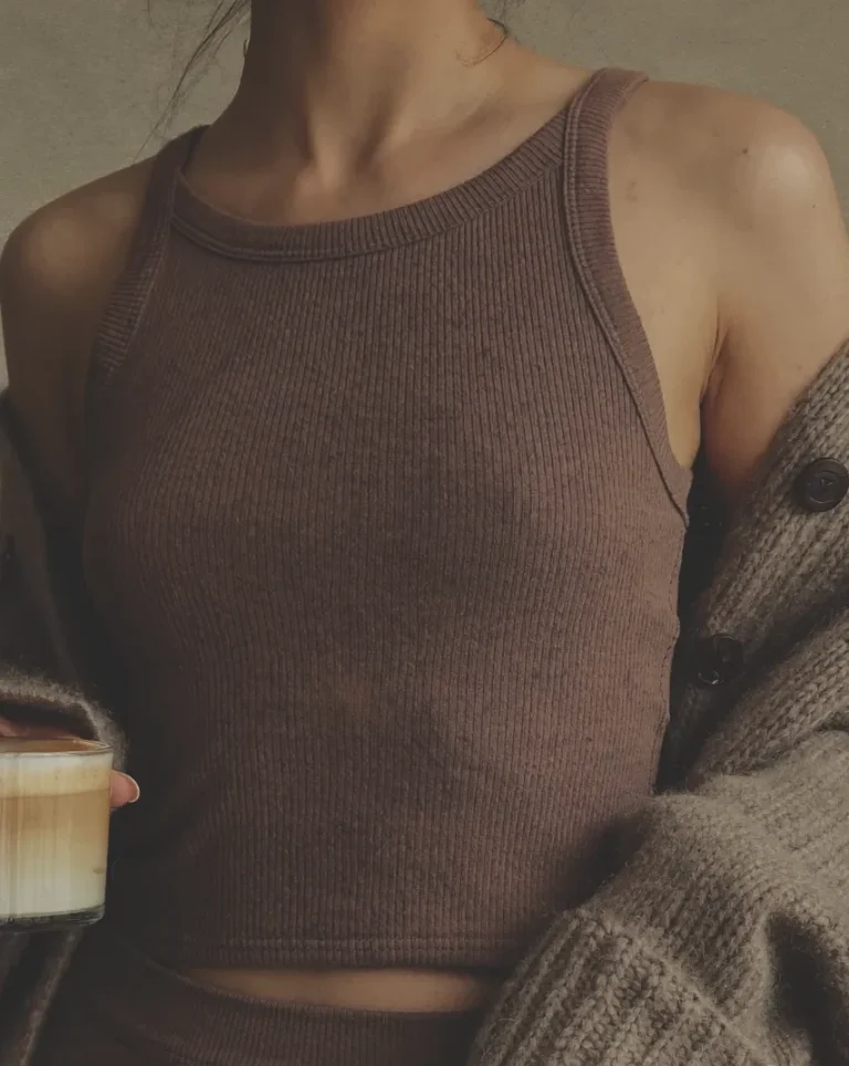 Person wearing a brown ribbed sleeveless top and a chunky cardigan draped over the shoulders, holding a glass mug of coffee.