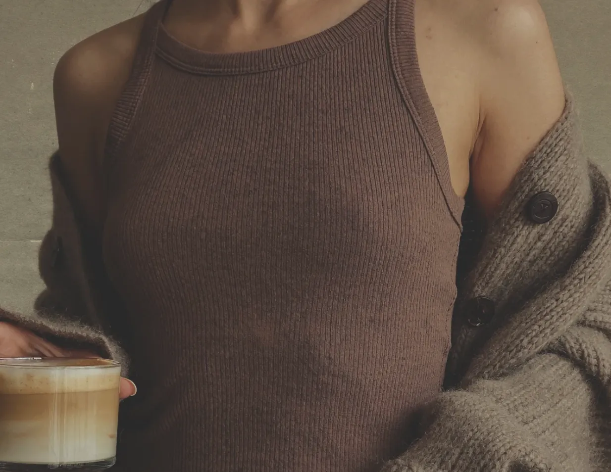 Close-up of an individual wearing a brown ribbed tank top and gray cardigan, holding a glass of layered latte.