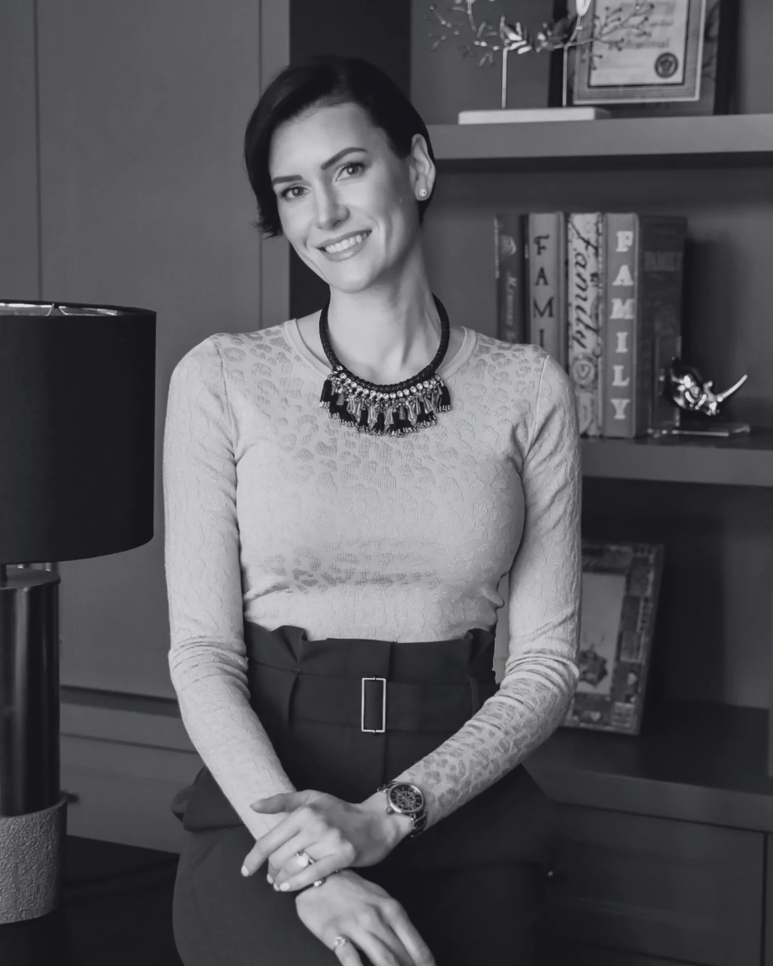 Smiling woman with short dark hair, wearing a lace top and statement necklace, seated at a desk in an office.