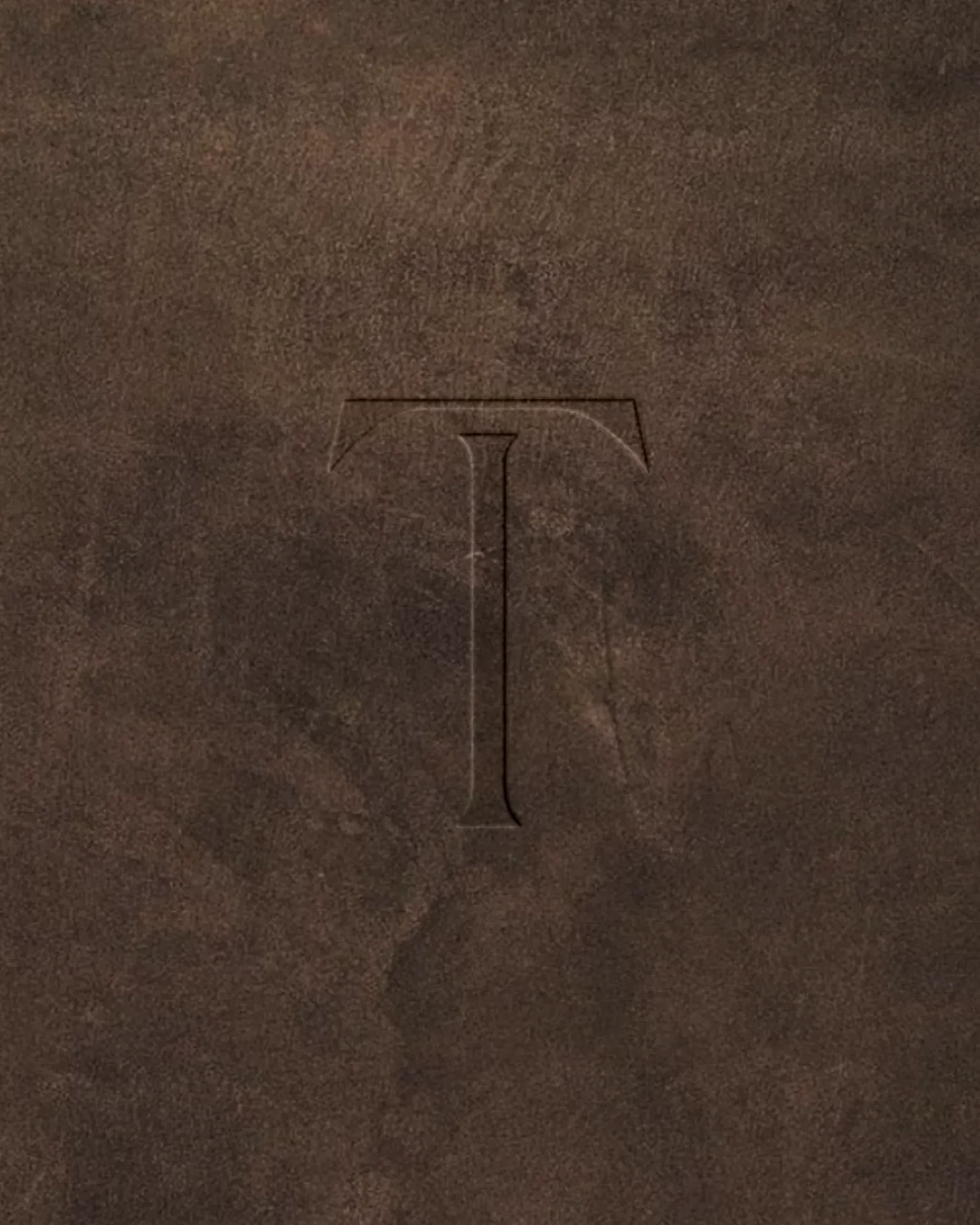 Embossed outline of a capital T inside a rounded-rectangle frame on a brown textured surface