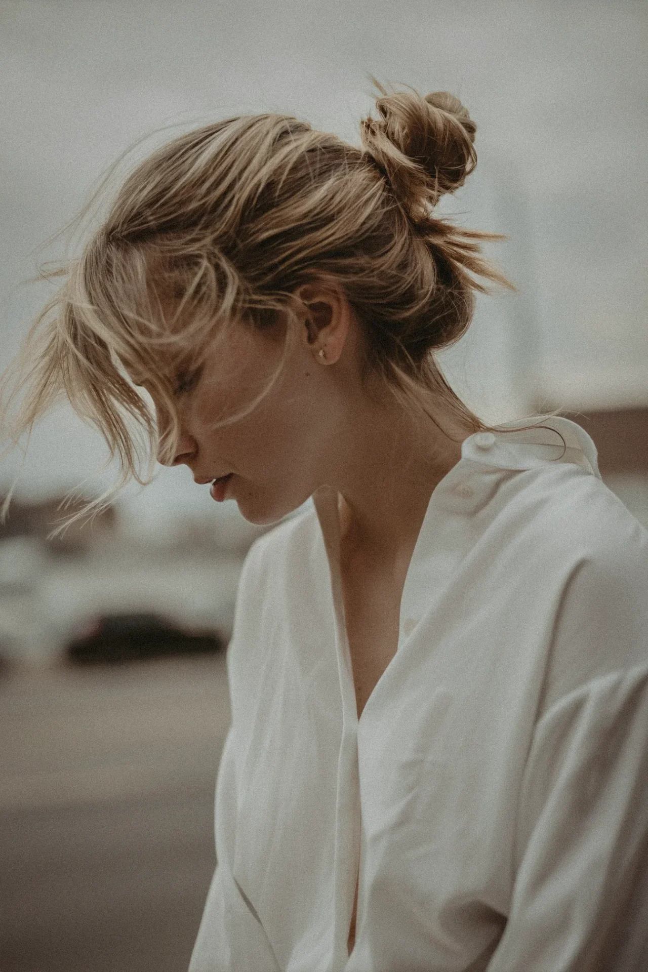 Profile of a woman with blond hair in a loose bun, wearing a white blouse, looking downward in a blurred outdoor setting.