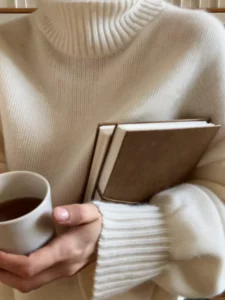 Person wearing a chunky cream sweater, holding two brown notebooks and a white mug of coffee close to their chest.