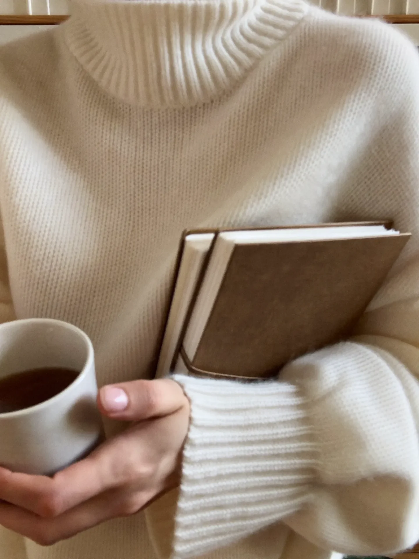 Person wearing a chunky cream sweater, holding two brown notebooks and a white mug of coffee close to their chest.