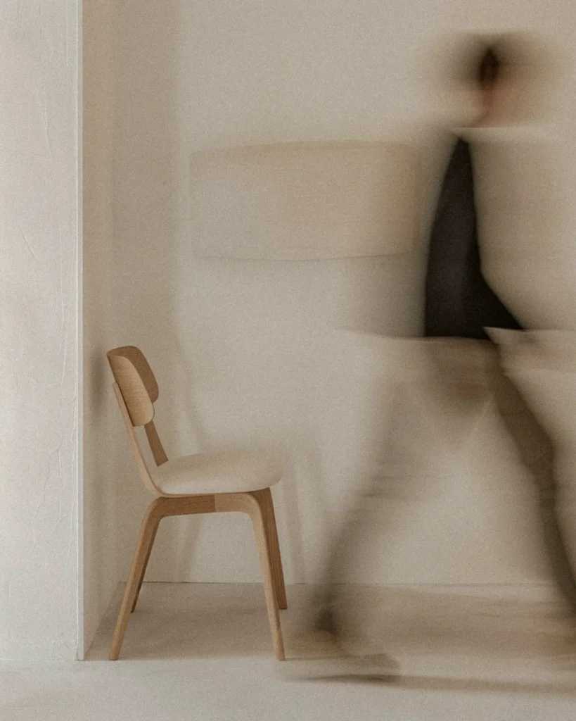 Wooden chair in a pale, minimal room; a blurred figure moves in the background, creating a sense of motion.