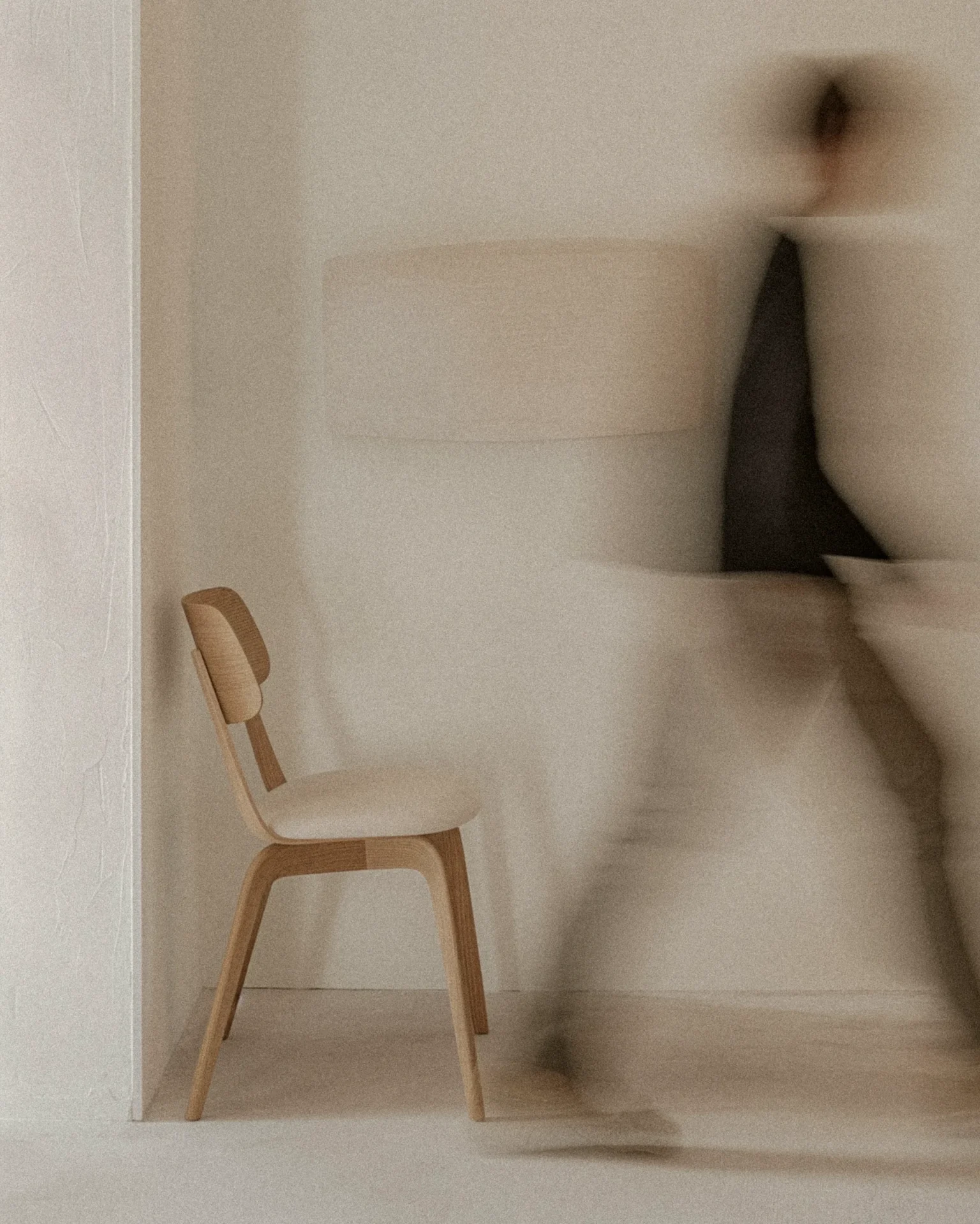 Wooden chair in a pale, minimal room; a blurred figure moves in the background, creating a sense of motion.