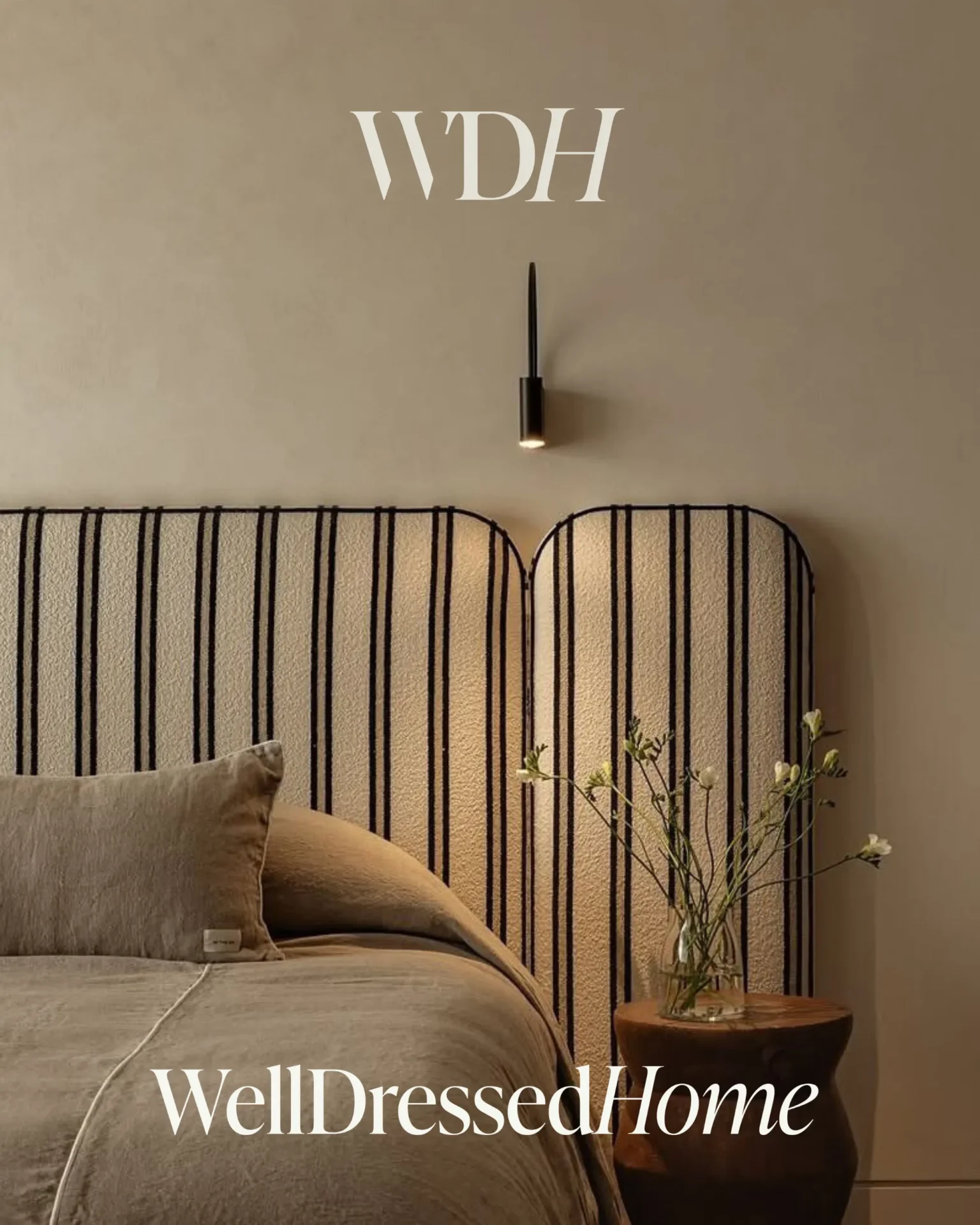 Bedroom with a vertically striped beige headboard, wall sconce above, and a round wooden side table holding a glass vase with branches; 'WellDressedHome' text at the bottom.