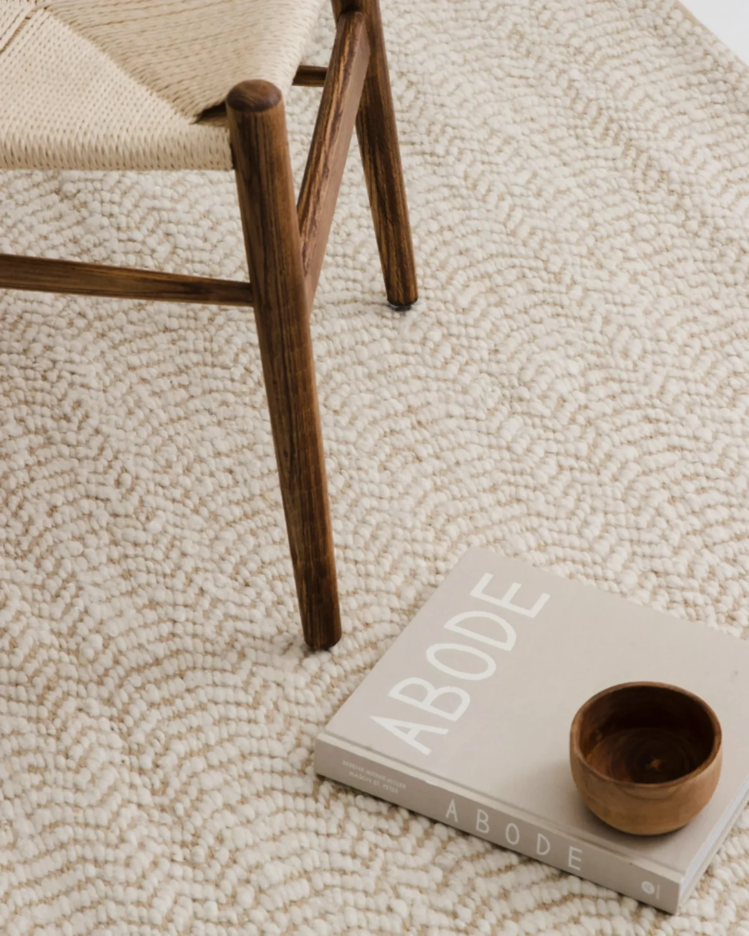 Close-up of a wooden chair leg resting on a cream textured rug, with a gray book titled 'ABODE' and two small wooden cups nearby.