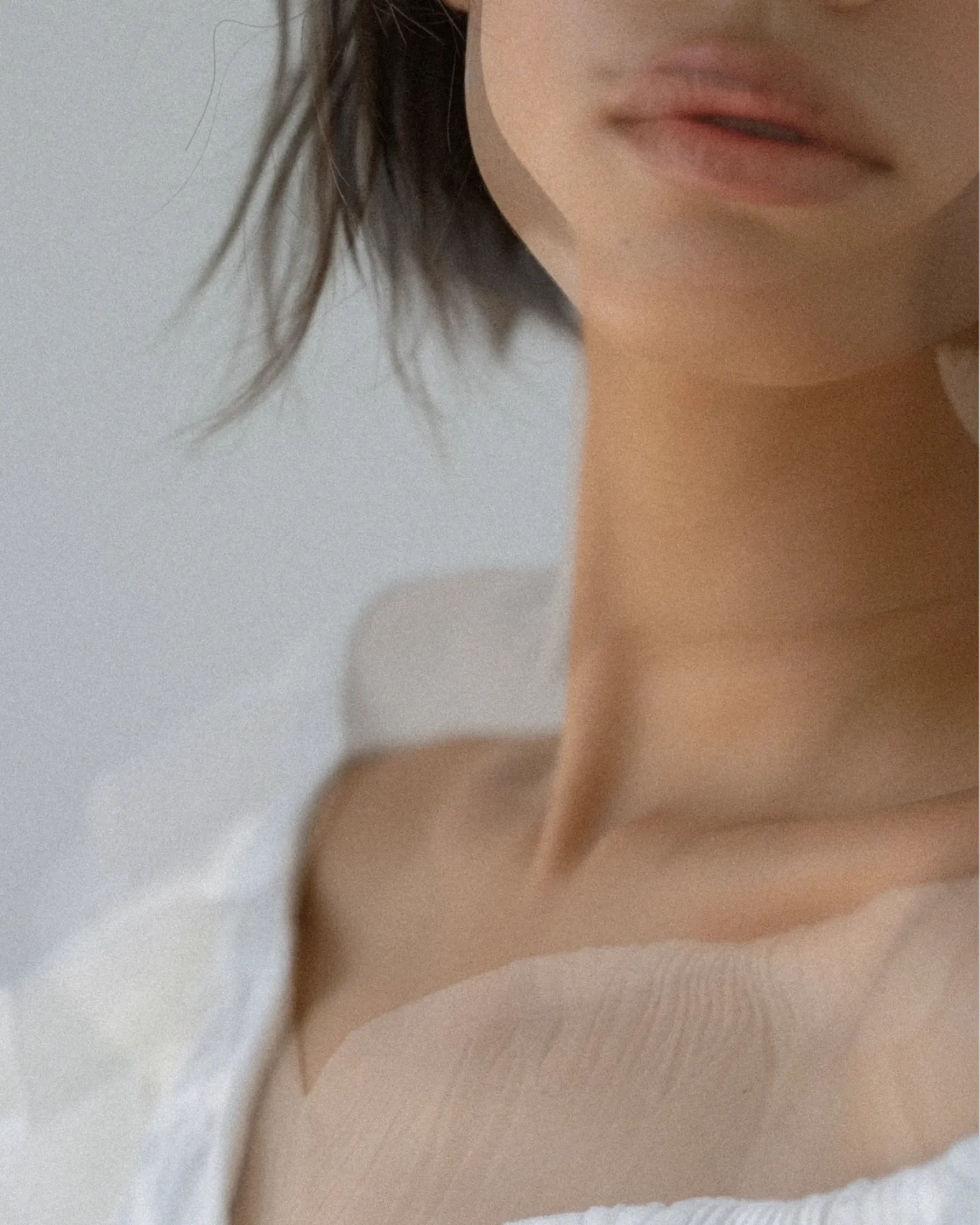 Close-up of a woman's lower face, neck, and shoulder in a soft-lit scene, wearing a white textured fabric top as the image is cropped at the mouth and neck.