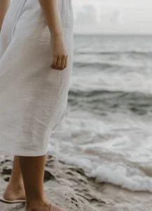 Close-up of a person in a white dress standing barefoot on the beach with the sea in the background.