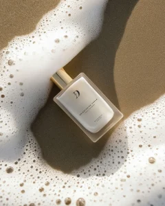 White rectangular bottle with a gold cap resting on wet sand beside white sea foam on a sunny beach.