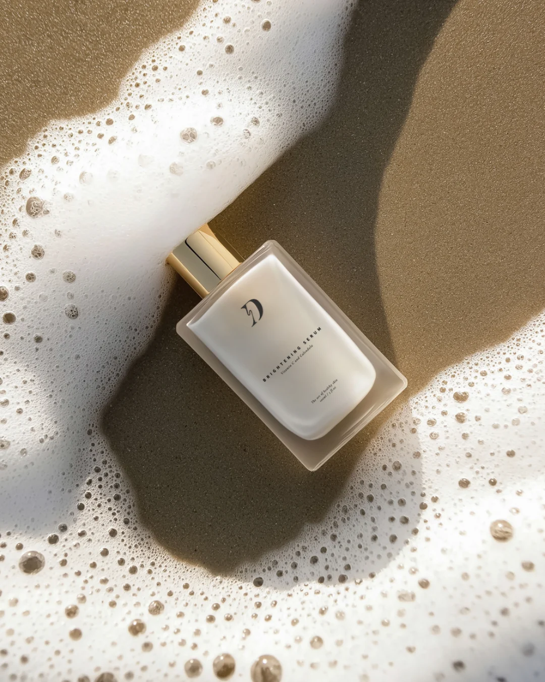 White rectangular bottle with a gold cap resting on wet sand beside white sea foam on a sunny beach.