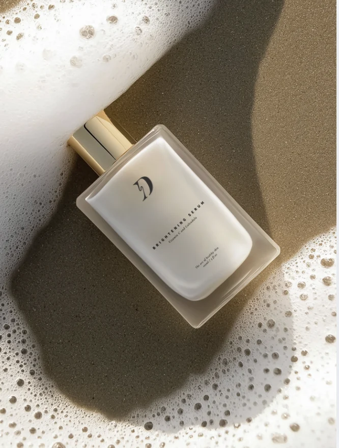 Frosted glass skincare bottle with a gold cap lies on wet sand with sea foam around it, labeled Brightening Serum.